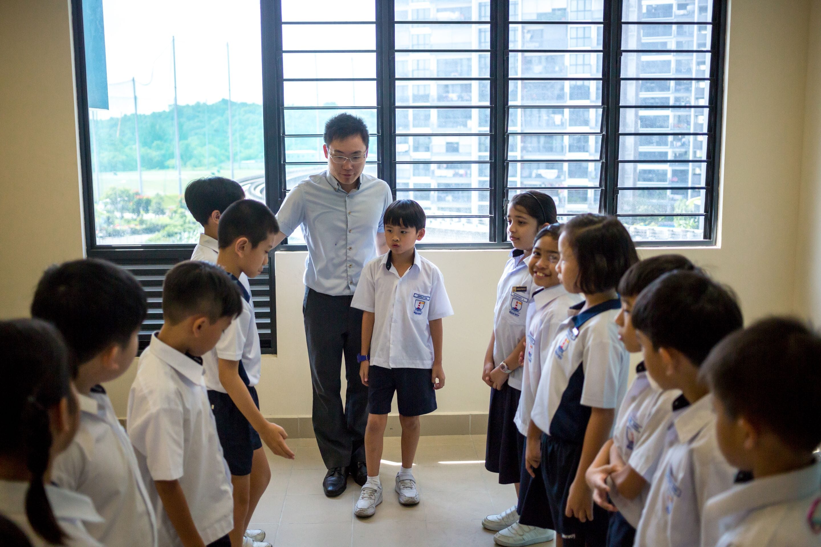 A male teacher in a blue shirt stands with a group of young students in white school uniforms indoors.